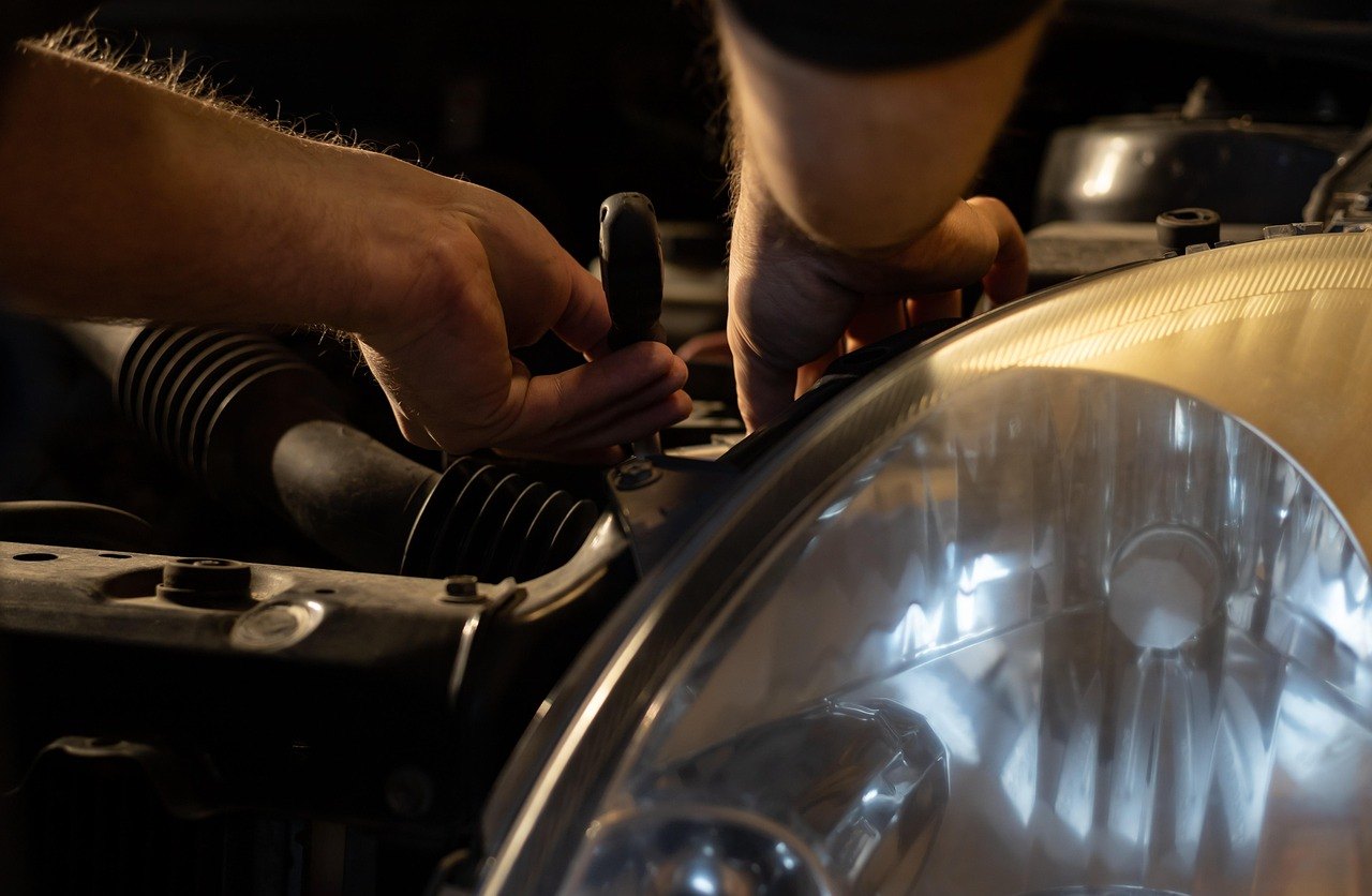A mechanic's hands using a tool in the engine bay of a car, beside the headlight
