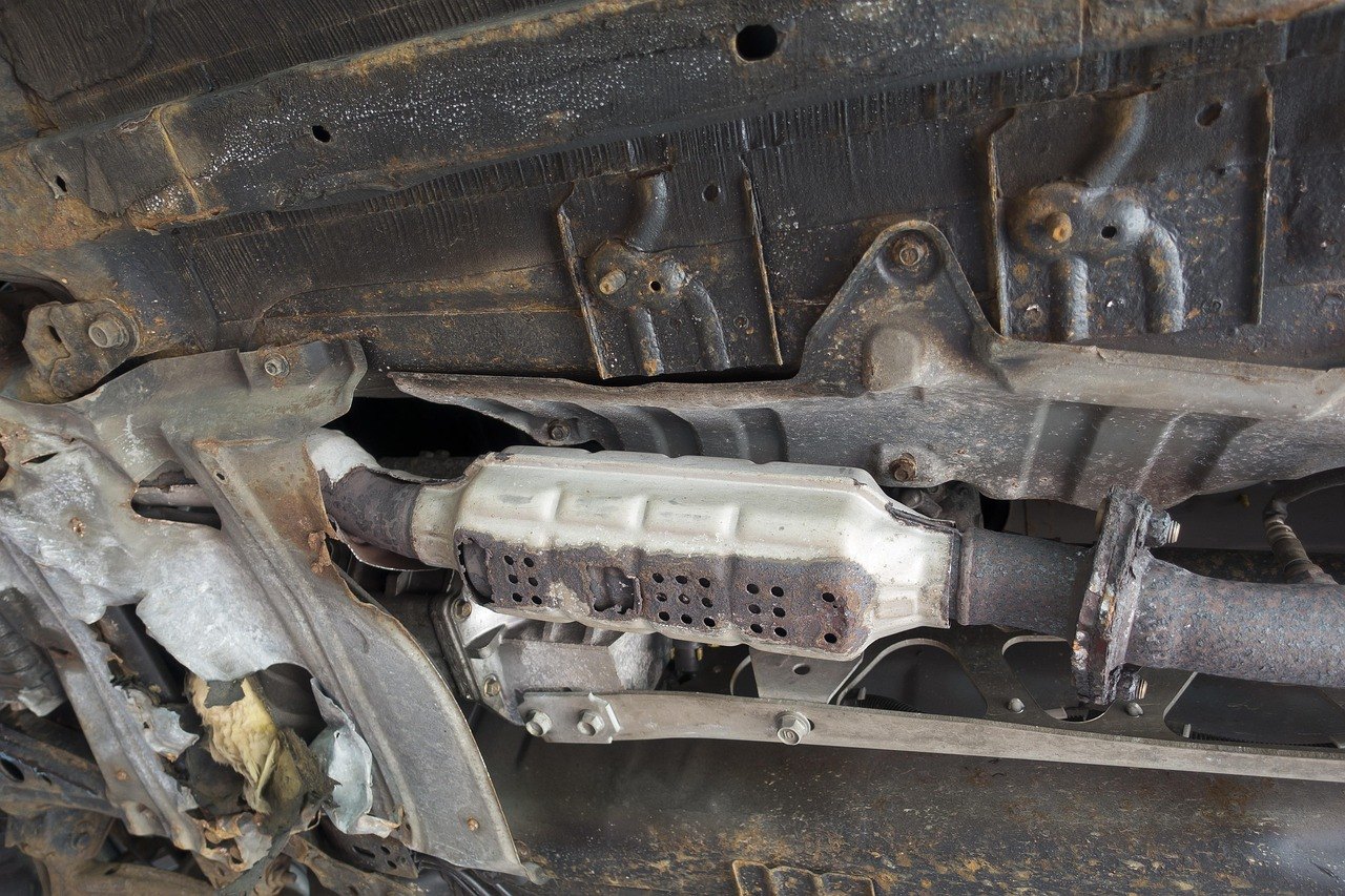 The underside of a car seen from below, showing the exhaust pipe and a catalytic converter with surface rust