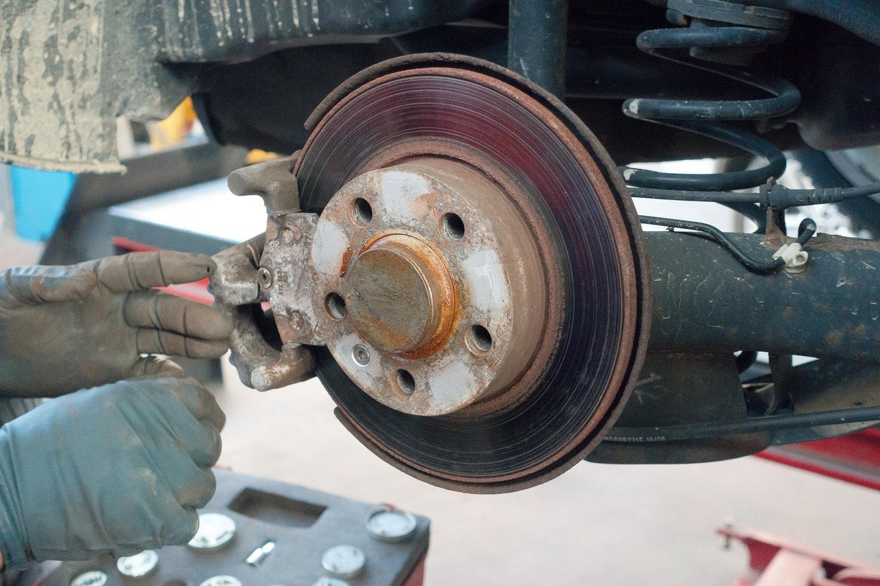 Gloved hands working at a brake disc with the wheel removed; a coil spring is visible behind it